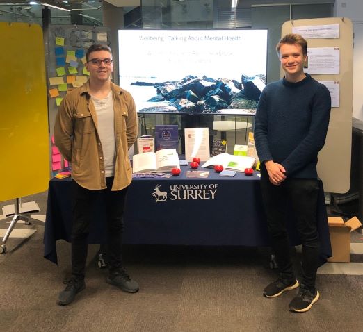Will and Aaron standing either side of the table in curator's corner, the table has a blue tablecloth with University of Surrey logo, and has some books displayed on it. In the background is a screen with a picture of some rocks and some boards with post it notes and paper pinned up.