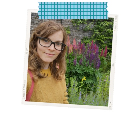 Daisy wearing a yellow sweater and glasses, standing in front of some plants.