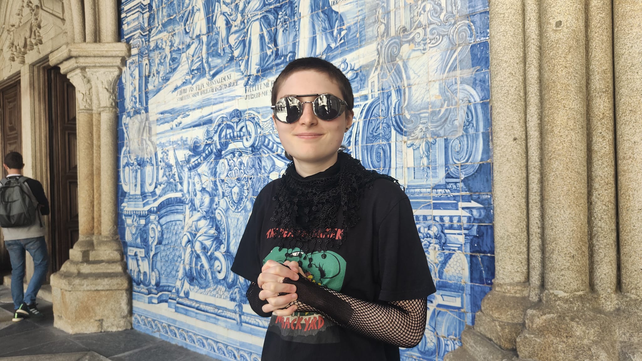 Giulia, wearing sunglasses and a black top, standing in front of blue tile picture and some stone columns.
